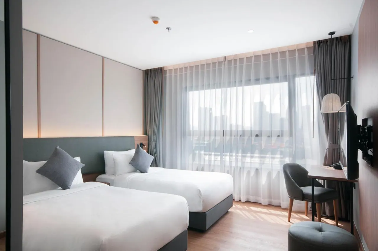 A twin bedroom with two beds dressed in white linen, a work desk with a chair and a wall-mounted television, and sheer curtains over a window showing city buildings.