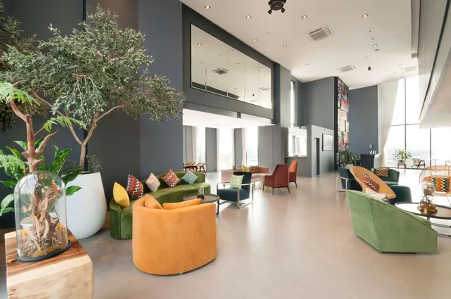 A lobby with green and orange sofas and armchairs, potted olive trees, and tall windows along the right wall.