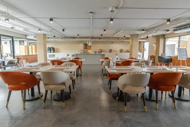 A dining room with orange and beige upholstered chairs at white-topped tables, with a buffet counter and chafing dishes in the background.