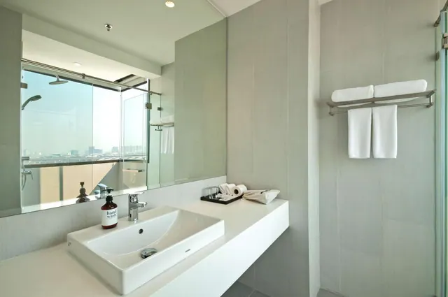A bathroom with a single white basin beneath a large mirror, a glass-walled shower, folded towels on a wall rail, and a hazy city skyline visible through the glass.