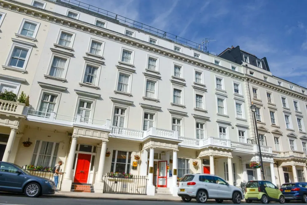 A white stucco terrace building with tall windows, balconies and red entrance doors, with parked cars and a pedestrian on the street in front.