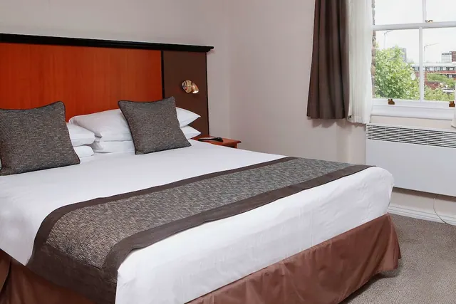 A double bed with white bedding, a brown runner and grey cushions sits beside a window looking out on trees and low buildings.