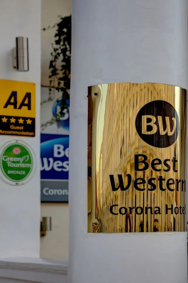 A brass Best Western plaque reading Corona Hotel is mounted on a white pillar beside an AA sign and a Green Tourism sticker.