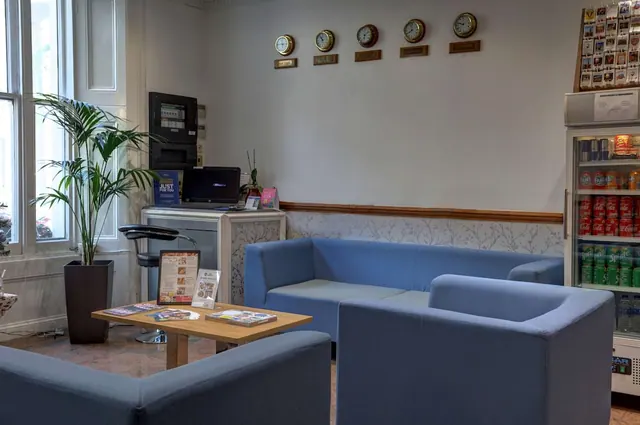 A lobby seating area with blue sofas, a low wooden table with magazines, a potted palm, wall clocks and a drinks fridge.