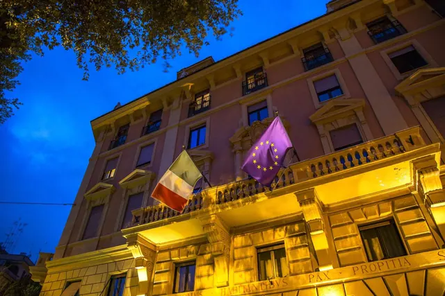 A multi-storey building facade lit at dusk, with an Italian flag and a European Union flag hanging from a balcony.