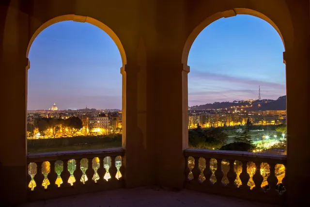 Two arched openings with stone balustrades overlook illuminated Rome at dusk, with the dome of St. Peter's Basilica visible on the left.