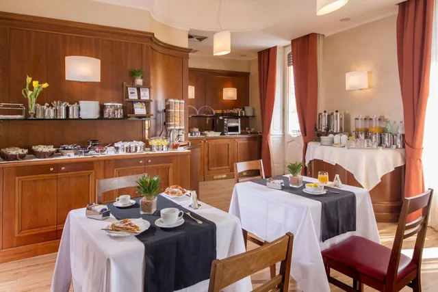 A breakfast dining room with set tables, a wooden sideboard holding pastries, cups and juice dispensers, and red curtains beside a window.