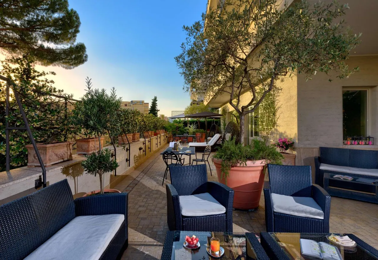 A furnished outdoor terrace with wicker seating, glass tables, olive trees and large terracotta planters under a clear sky.
