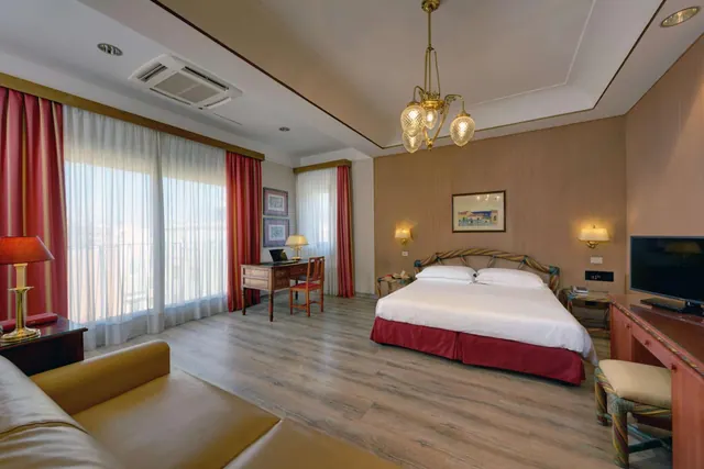 A hotel bedroom with a king bed, a wooden desk and chair by the windows, a television on a dresser and a leather sofa in the foreground.