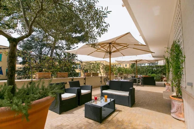 A covered outdoor terrace with black wicker armchairs and a sofa, a glass coffee table with two drinks, cream parasols, and potted olive trees and plants along the edge.