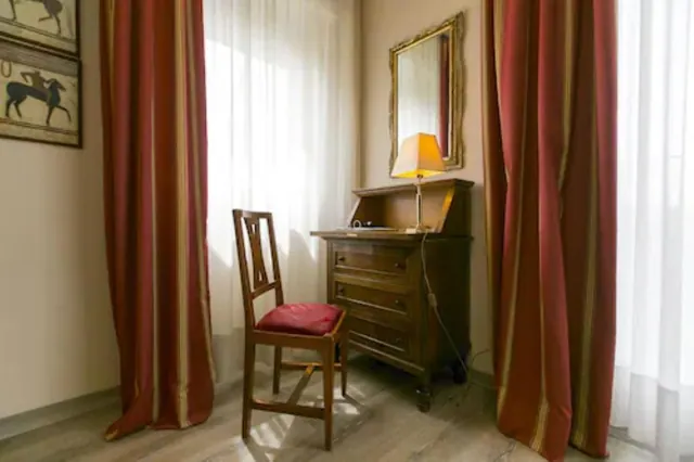 A wooden chair with a red seat stands beside a wooden secretaire with a lamp and mirror, between red curtains and sheer white curtains.