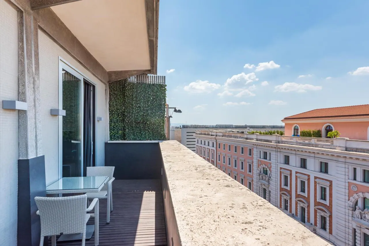 A private balcony with a glass table and two white wicker chairs overlooks neighbouring ochre and pink buildings and the city under a blue sky.