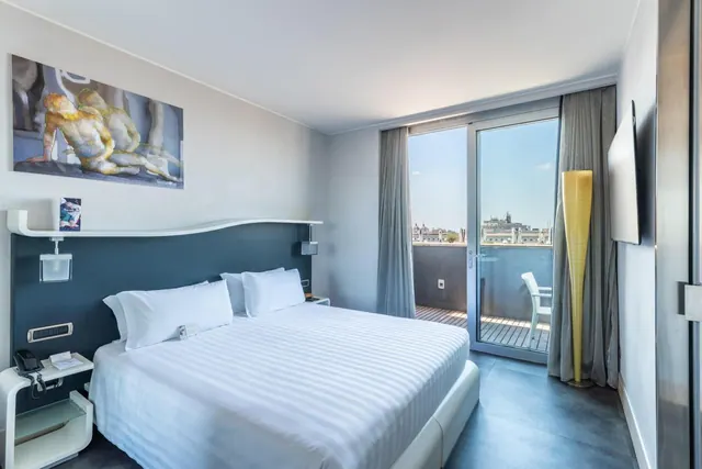A bedroom with a made king bed, a large painting above the headboard, and a glass door opening onto a balcony with rooftop views.