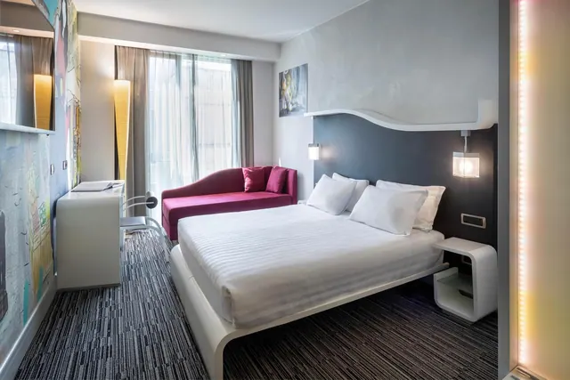 A bedroom with a double bed, a magenta sofa beside tall sheer-curtained windows, and a white desk against a colourful wall.
