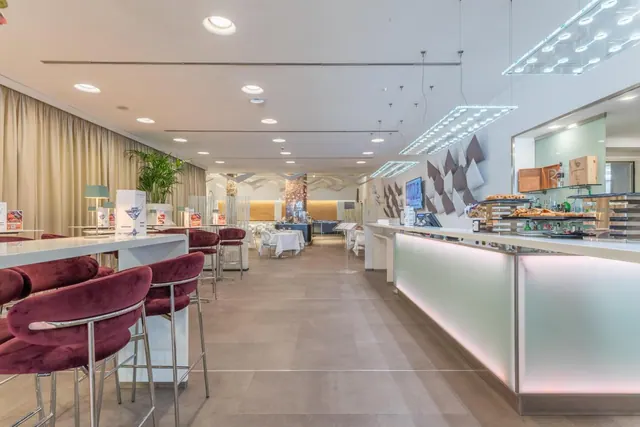 A hotel bar with burgundy velvet stools at a white counter, a backlit pastry counter on the right, and a dining room with set tables in the background.