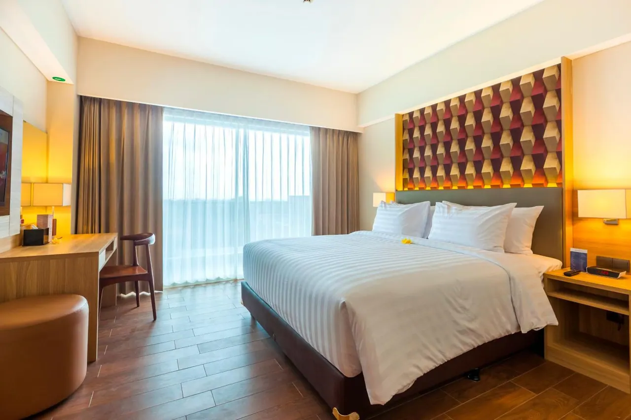 A king bed with white striped linens sits beside a wooden desk and a tall window with sheer curtains in a hotel bedroom.