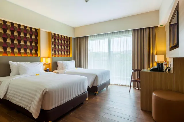 A twin bedroom with two beds with white striped linen, a wall-mounted television, a wooden desk with a kettle, and a large window with sheer curtains looking onto greenery.