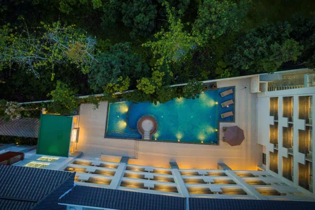 Aerial dusk view of a rectangular swimming pool with a circular centre feature, sun loungers and a parasol beside it, surrounded by trees and a hotel building.