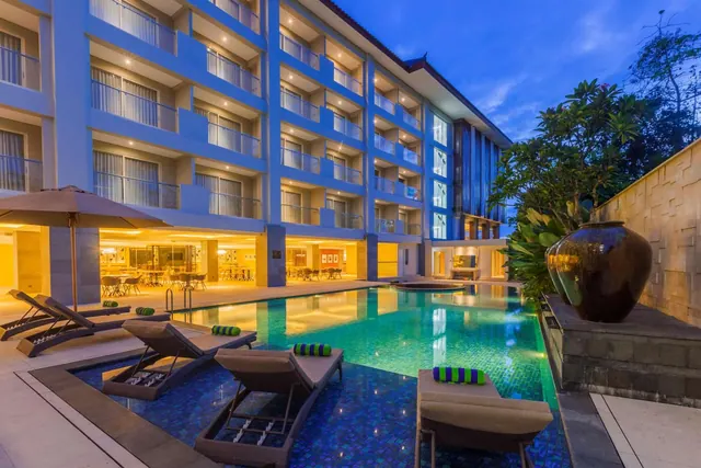 Evening view of an outdoor swimming pool with loungers and a parasol beside a multi-storey hotel building with balconies.