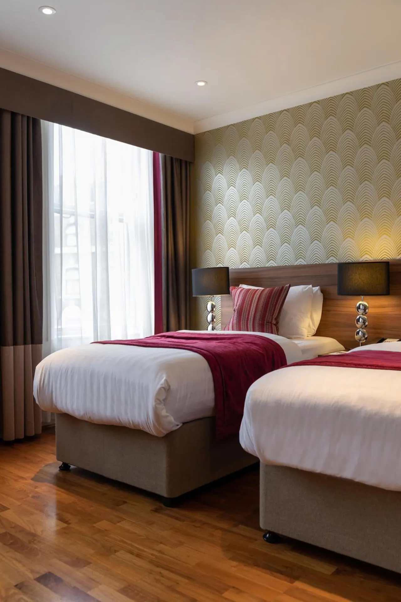 Two single beds with red throws sit beside a window with sheer curtains and a wall covered in patterned wallpaper.