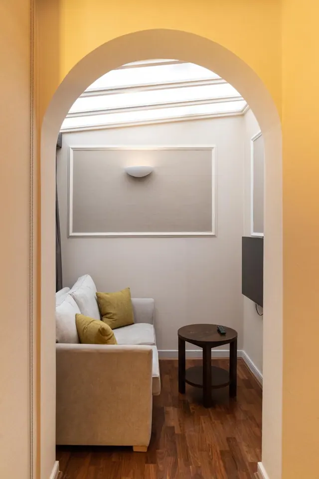 A seating area with a beige sofa, yellow cushions and a round dark wood side table is seen through an arched doorway, with a skylight above and a wall-mounted television on the right.