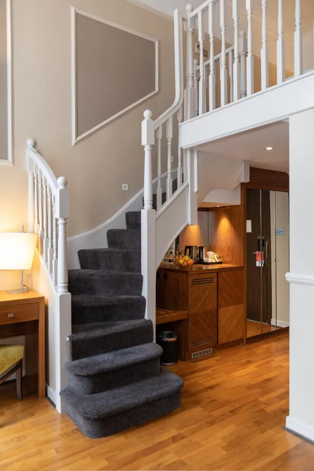 A staircase with grey carpeted treads rises to a white-railed mezzanine, beside a kettle and fruit station and a lift door.