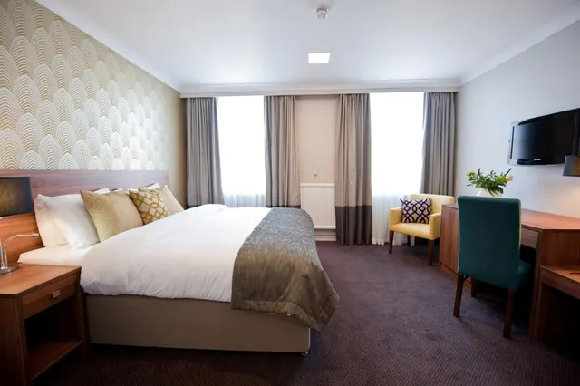A made-up double bed with a patterned gold feature wall, a yellow armchair, a desk with a chair and a wall-mounted television in a hotel bedroom.