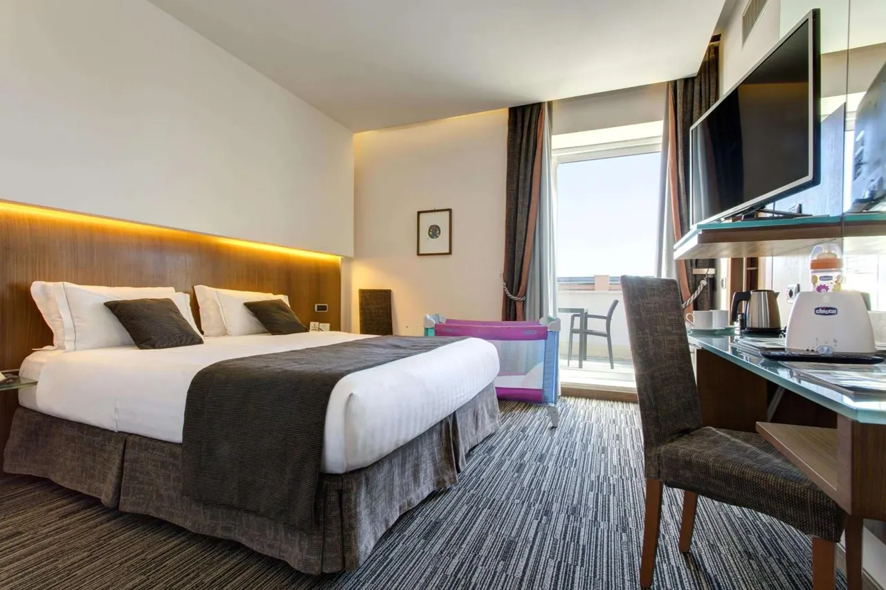 A double bed with a lit wooden headboard, a travel cot, a glass desk with a wall-mounted television and kettle, and a glass door onto a balcony with a table and chairs.