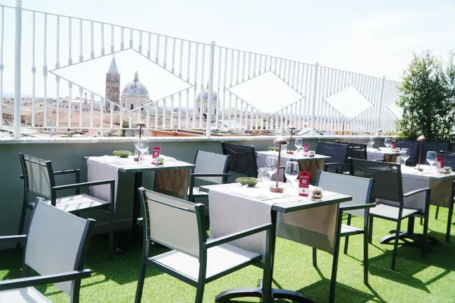 A rooftop terrace with laid dining tables and chairs on artificial turf, with domes and a brick bell tower of Rome visible beyond the white railing.