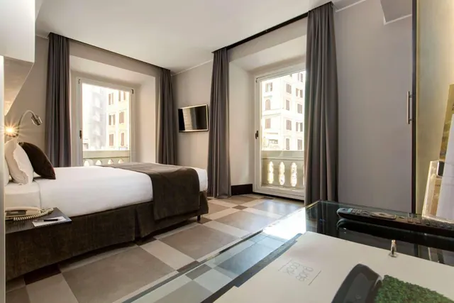 A double bed sits beside a wall-mounted television, with two tall windows opening onto a small balcony and neighbouring buildings.