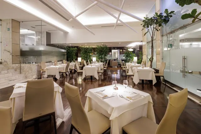 A dining room with white-clothed tables, beige leather chairs, a dark wood floor and potted plants, with a bar area visible in the background.