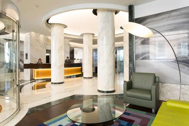 The lobby has tall marble columns, a reception desk with two staff members behind it, a green leather armchair, a glass coffee table and a yellow sofa.