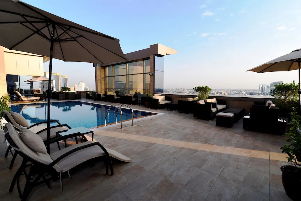 Rooftop swimming pool with sun loungers, shade umbrellas and lounge seating, with a hazy city skyline in the background.