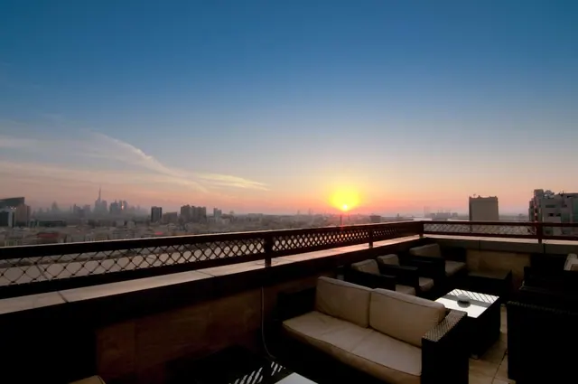 A rooftop terrace with cream cushioned sofas and wicker lounge chairs overlooks a Dubai skyline at sunset.
