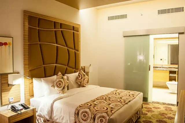 A hotel bedroom with a double bed under a large upholstered headboard and a bedside telephone, with a sliding frosted glass door open to an en-suite bathroom containing a toilet and vanity.
