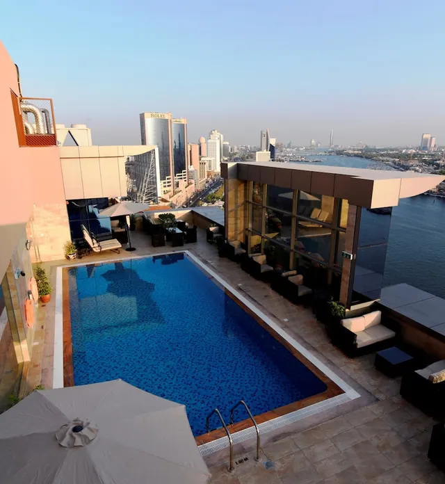 A rooftop swimming pool with loungers, umbrellas and seating, overlooking city towers and a waterway under a clear sky.