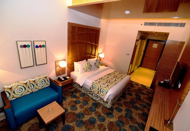 A hotel bedroom with a double bed, a blue sofa with patterned cushions, framed prints on the wall, and a wall-mounted television.