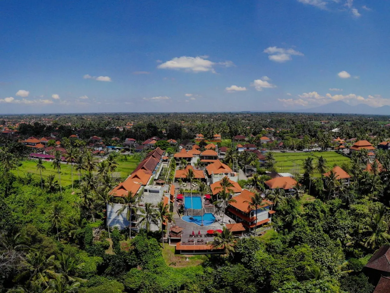 Aerial view of a resort with orange-roofed buildings, a swimming pool with red umbrellas and sun loungers, surrounded by palm trees and rice fields.