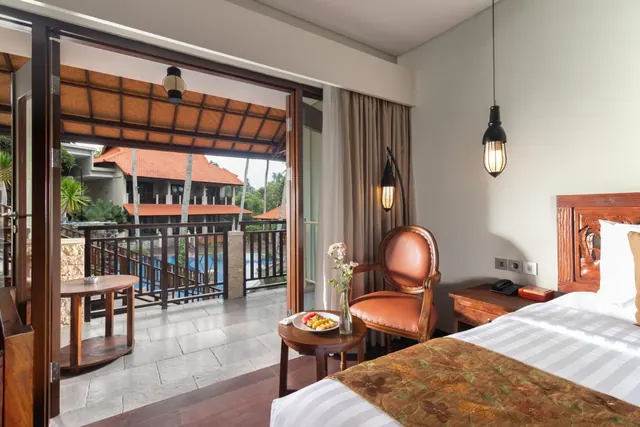A bedroom with a double bed, an armchair and a side table with a fruit plate, with sliding doors open onto a balcony overlooking a pool and trees.