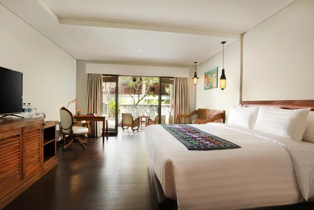 A bedroom with a large bed, a wooden cabinet with a wall-mounted television, a writing desk and chair, and sliding glass doors opening onto a balcony overlooking a garden.