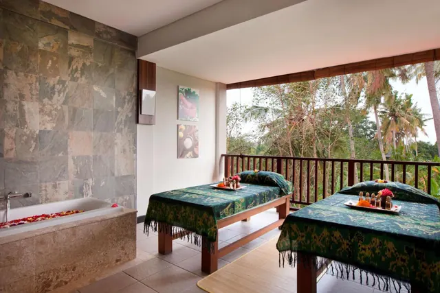A spa treatment room with two massage tables with green batik covers, a stone-tiled bathtub with flower petals, and an open side looking onto palm trees.