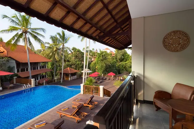 A covered balcony overlooks a swimming pool with sun loungers, red parasols and palm trees beyond.