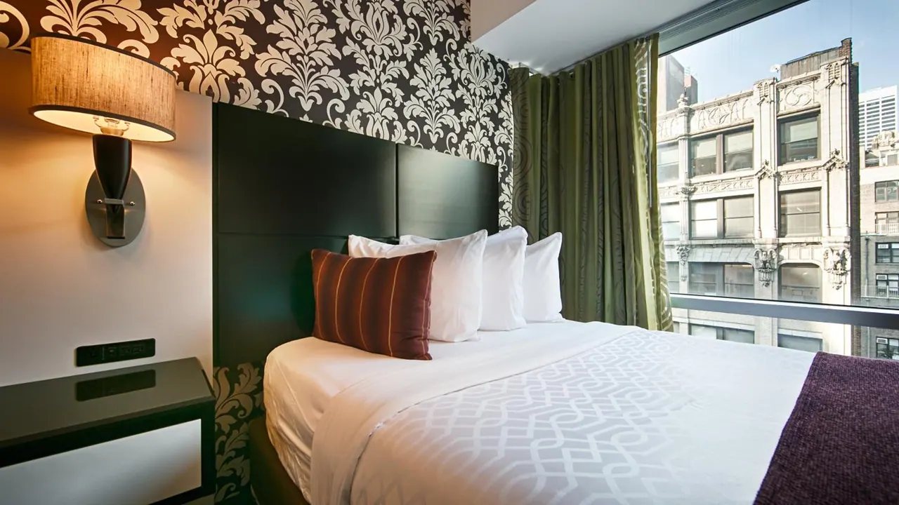 A made double bed with white pillows and a brown striped cushion sits beside a large window that looks onto an ornate stone building across the street, with a damask-patterned wall and a wall lamp.