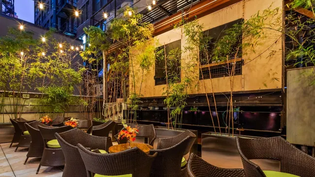 An outdoor terrace at dusk with wicker chairs and green cushions, bamboo planting, string lights overhead and a flower arrangement on a round table.