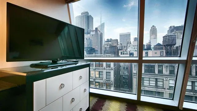 A flat-screen television sits on a white-drawered dresser beside floor-to-ceiling windows overlooking a city skyline with tall buildings and rooftop water towers.