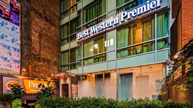Exterior of a Best Western Premier hotel building at dusk, with lit windows, green curtains and a hedge in the foreground.
