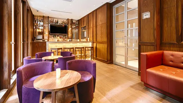 A wood-panelled bar lounge with purple velvet armchairs, round wooden tables with candles, a backlit bar with stools, a wall-mounted television and an orange leather sofa.