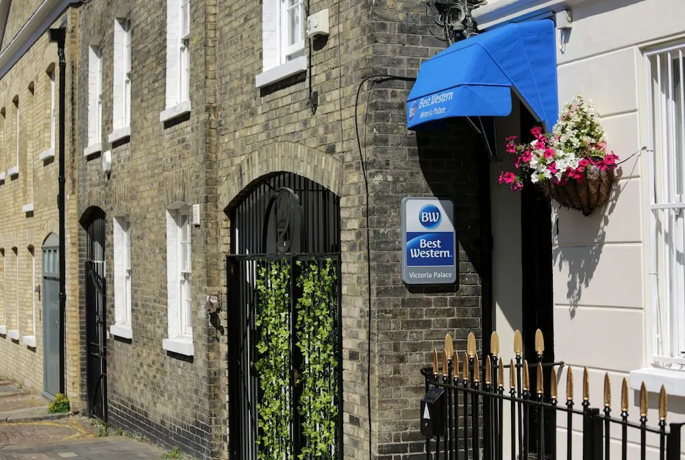The street-facing entrance of a brick hotel building shows a blue awning, a Best Western sign, a gated doorway with ivy, a hanging flower basket and a black iron railing.