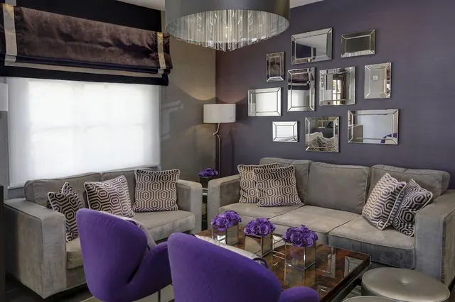 A living area with grey sofas, purple swivel chairs, a wall of mirrors, a drum chandelier and a coffee table holding purple flowers in glass vases.