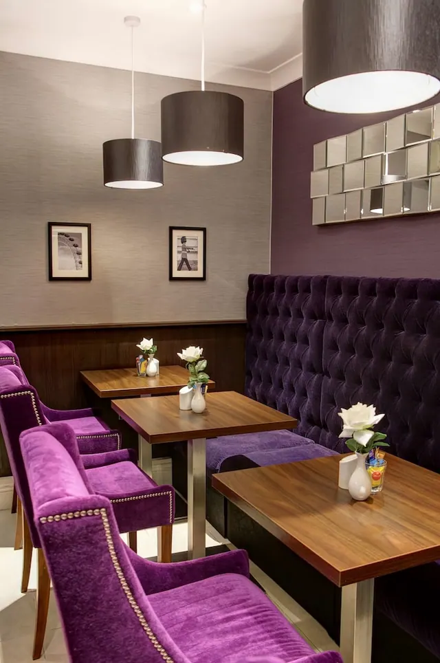 A dining area with purple velvet chairs and a tufted purple banquette, wooden tables holding white roses in vases, and pendant lamps overhead.